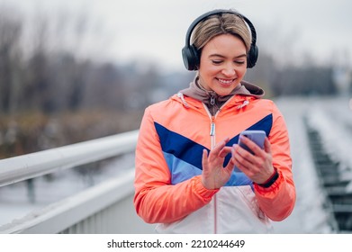 Happy Beautiful Woman In Sportswear Using Smartphone During Training On The Bridge At Winter. Healthy Lifestyle, Winter Fitness, Cold Weather.