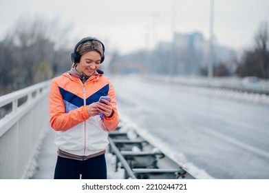 Happy Beautiful Woman In Sportswear Using Smartphone During Training On The Bridge At Winter. Healthy Lifestyle, Winter Fitness, Cold Weather.