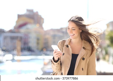 Happy beautiful woman walking and writing or reading sms messages on line on a smart phone while the wind moves her hair in a street of a port - Powered by Shutterstock