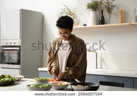 Happy beautiful young 30s African American woman in eyeglasses chopping fresh vegetables for salad on countertop, enjoying cooking vegan food in kitchen, healthcare hobby activity, dieting concept.