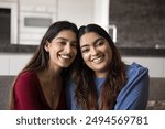 Happy beautiful young adult Indian sibling sisters sitting together on couch, hugging with love, looking at camera with toothy smiles, enjoying friendship, good close relationships. Family portrait