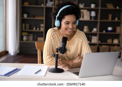 Happy beautiful young African ethnicity female radio program host or blogger in bright headphones recording audio or voice acting using stand microphone, streaming online of holding video call.