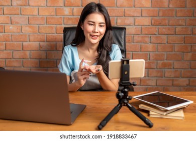 Happy beautiful young Asian woman using hand sanitizer gel on her hands while talking on video call  through the smart phone in her home office, new normal concept - Powered by Shutterstock