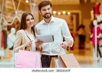 Happy beautiful young couple holding shopping bags, using a tablet and smiling while standing in mall - Powered by Shutterstock