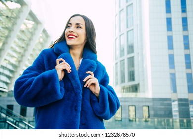 Happy Beautiful Young Cute Woman In Blue Vegan And Faux Fur Coat Is Posing On Urban City Background. Fashion Trendy Style Clothes