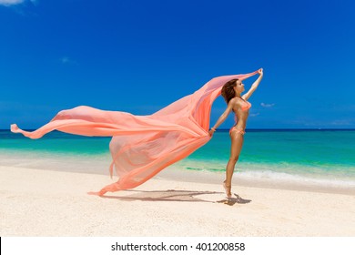 Happy beautiful young girl in pink bikini with waving in the wind pink fabric on a tropical beach. Blue sea in the background. Summer vacation concept.