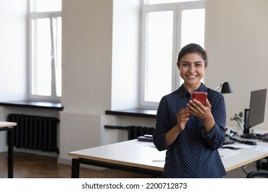 Happy Beautiful Young Indian Business Woman Holding Mobile Phone, Standing At Work Table, Looking At Camera, Smiling. Cheerful Office Employee Girl Using Online App On Smartphone. Head Shot Portrait