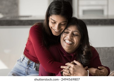 Happy beautiful young Indian daughter woman embracing senior mom from behind with love, tenderness. Cheerful mother and adult kid hugging with closed eyes on home couch, smiling, laughing - Powered by Shutterstock