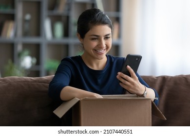 Happy Beautiful Young Indian Woman Unboxing Carton Parcel At Home, Checking Order In Cellphone Shopping Application, Leaving Positive Review, Feeling Satisfied With Product Or Quick Delivery Service.