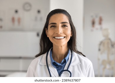 Happy beautiful young Latin doctor woman in white uniform coat and stethoscope posing in clinic office, looking at camera with perfect toothy smile, laughing. Healthcare medical professional portrait - Powered by Shutterstock