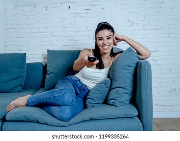 Happy beautiful young latin woman having fun watching TV with control remote in hand sitting on couch at home in television relax lifestyle concept. - Shutterstock ID 1199335264