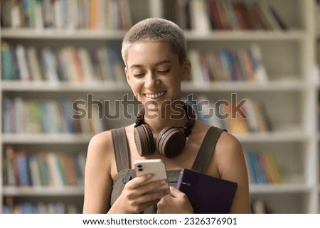 Happy beautiful young student girl with trendy short haircut typing message on mobile phone, making call, using learning app in campus library, smiling, laughing, enjoying Internet communication