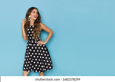 Happy beautiful young woman in black cocktail dress is holding hand on hip, looking away and talking. Three quarter length studio shot on blue background. - Shutterstock ID 1682378299