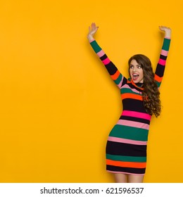 Happy Beautiful Young Woman In Colorful Vibrant Striped Dress Is Holding Arms Raised, Shouting And Looking Away. Three Quarter Length Studio Shot On Yellow Background.