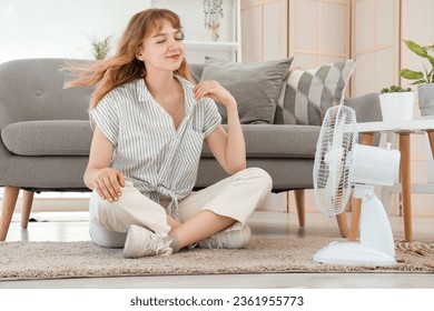 Feliz y bella joven con ventilador eléctrico sentado en el suelo en la sala de estar