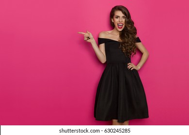 Happy beautiful young woman in elegant black cocktail dress is shouting, looking at camera and pointing. Three quarter length studio shot on pink background.