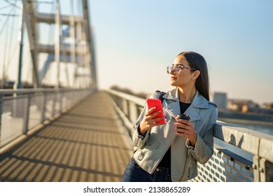 Happy beautiful young woman enjoying in sunset, she's walking on city bridge while holding phone and cup of coffee and posing for photo shoot. - Shutterstock ID 2138865289