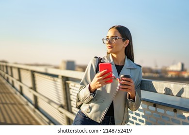 Happy beautiful young woman enjoying in sunset, she's walking on city bridge while holding phone and cup of coffee and posing for photo shoot. - Shutterstock ID 2148752419