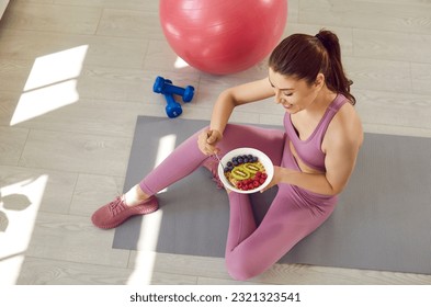 Happy beautiful young woman having good healthy snack after workout, sitting on yoga mat on floor with fit ball and dumbbells, eating muesli, kiwi, blueberries and raspberries. Sport food diet concept - Powered by Shutterstock