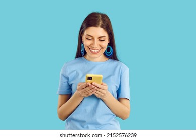 Happy beautiful young woman looking at her mobile phone and smiling. Pretty girl in casual T shirt standing on blue background, holding cell phone and reading funny message she received from boyfriend
