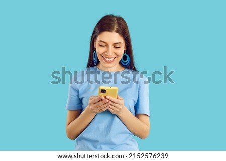 Happy beautiful young woman looking at her mobile phone and smiling. Pretty girl in casual T shirt standing on blue background, holding cell phone and reading funny message she received from boyfriend