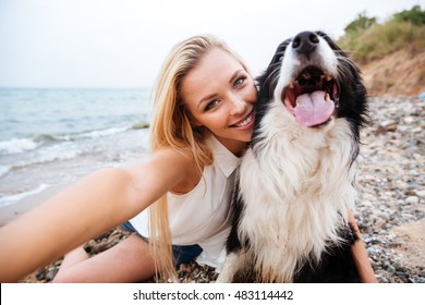 Happy Beautiful Young Woman Sitting And Making Selfie With Dog On The Beach