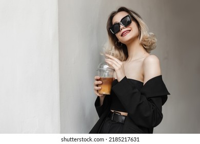 Happy Beautiful Young Woman With A Smile And Sunglasses Wearing Fashionable Black Elegant Clothes Walking On The Street Near The White Wall And Drinking Cold Coffee. Girl Drinks And Enjoy 