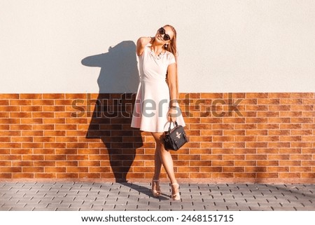 Happy beautiful young woman with a smile with vintage sunglasses in a pink summer dress with a bag with heels stands near a wall on a sunny day