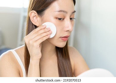 Happy beauty, beautiful asian young woman, girl smiling, looking in mirror, holding cotton pad, applying lotion by wipe on her face, removing makeup before shower at home. Skin care routine people.