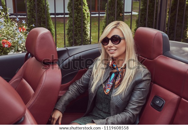 Happy beauty blonde female in sunglasses\
wearing green dress and gray leather jacket sitting on a back seat\
in a convertible car on summer\
vacation.