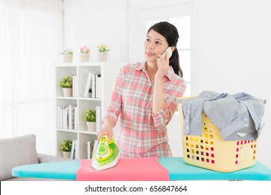 Happy Beauty Business Woman Using Mobile Cell Phone Calling For Family When She Ironing Messy Clothing In Living Room At Home. Housewife Doing Housekeeping Concept.