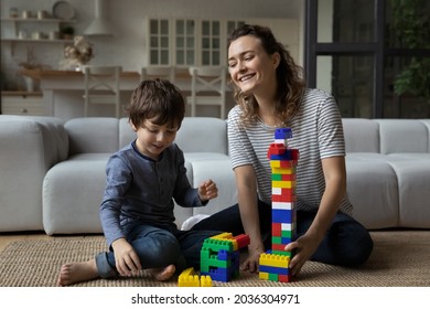 Happy best mom helping cute toddler son to build high toy tower from plastic construction blocks, playing with kid on carpet and heating floor at home, relaxing, enjoying leisure and motherhood
