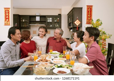 Happy Big Asian Family Sitting At Dinner Table And Celebrating Lunar New Year, Couplets With Best Wishes For Coming Year In The Background