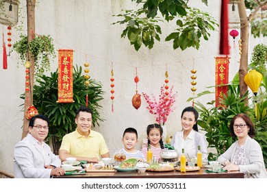 Happy Big Asian Family Sitting At Big Dinner Table Outdoors And Celebrating Lunar New Year, Decorations With Best Wishes Inscription In Background