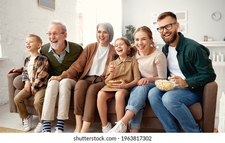 Happy Big Family Enjoying Weekend Together At Home. Grandparents, Mother, Father And Two Little Kids, Sister And Brother, Sitting On Sofa And Eating Popcorn While Watching Funny Movie Comedy On TV