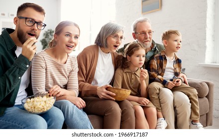 Happy Big Family Enjoying Weekend Together At Home. Grandparents, Mother, Father And Two Little Kids, Sister And Brother On Sofa And Eating Popcorn While Watching With Surprise Movie Comedy On TV