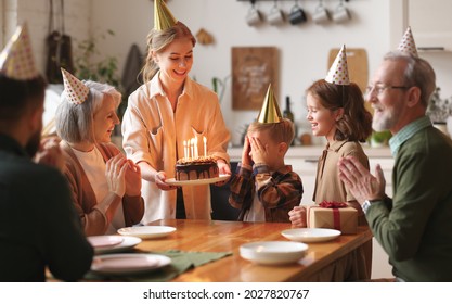 Happy big family gathered in living room table to celebrate birthday of cute little boy, excited child covering eyes with hands while mother surprising her by bringing chocolate cake with lit candles - Powered by Shutterstock