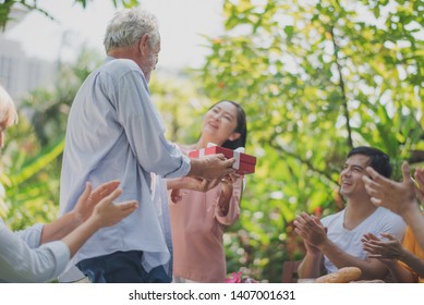 Happy Big Family Have A Lunch At Outdoor In Green Garden. Grand Father's Birthday Party On Picnic Table In Summer. The Asian Woman Give Gifts To Old Europe Man. Big Family Outdoor Lunch Party Concept.