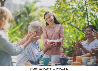 Happy Big Family Have A Lunch At Outdoor In Green Garden. Grand Father's Birthday Party On Picnic Table In Summer. The Asian Woman Give Gifts To Old Europe Man. Big Family Outdoor Lunch Party Concept.