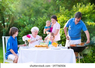 Happy Big Family - Young Mother And Father With Kids, Teen Age Son, Cute Toddler Daughter And A Little Baby, Enjoying BBQ Lunch With Grandmother Eating Grilled Meat In The Garden With Salad And Bread