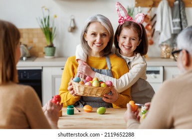 Happy Big Multi-generation Family Grandparents, Parents And Child Prepare For Easter Holiday At Home Together, Grandmother And Granddaughter In Bunny Ears Hugging While Painting Eggs In Kitchen