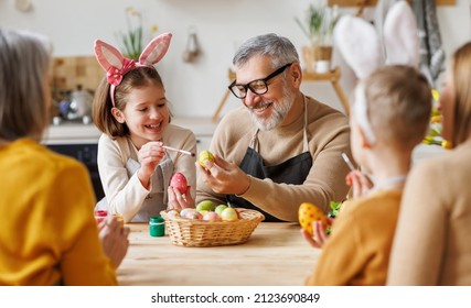 Happy Big Multi-generation Family Grandparents, Parents And Children Prepare For Easter Holiday At Home Together, Grandfather And Granddaughter In Bunny Ears Hugging While Painting Eggs In Kitchen