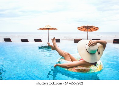 Happy Bikini Asian Woman With Hat Relax On Pool Float In Swimming Pool Against Beach Chair And Sea Background At Day, Summer Vacation Concept