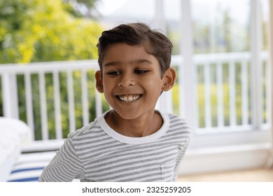 Happy biracial boy smiling in bedroom in front of window with view of garden. Childhood, wellbeing, domestic life and lifestyle, unaltered. - Powered by Shutterstock