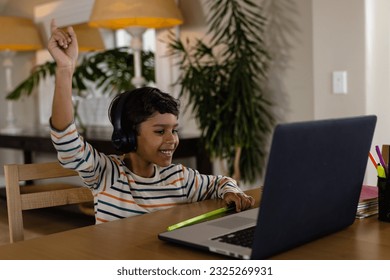Happy biracial boy using headphones and laptop for online lesson at home, raising hand. Education, learning, childhood, communication, technology and domestic life, unaltered. - Powered by Shutterstock