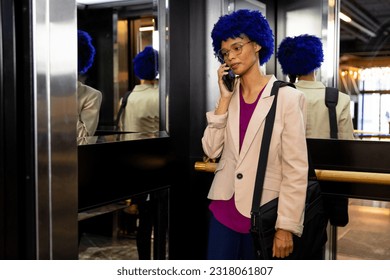 Happy biracial casual businesswoman with blue afro talking on smartphone in office elevator. Casual office, business, communication and work, unaltered. - Powered by Shutterstock