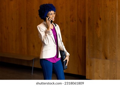 Happy biracial casual businesswoman with blue afro talking on smartphone in office corridor. Casual office, business, communication and work, unaltered. - Powered by Shutterstock