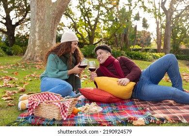 Happy biracial couple having picnic sitting on a rug and drinking wine in autumn garden and smiling. Inclusivity, domestic life, leisure time, romance and togetherness concept.