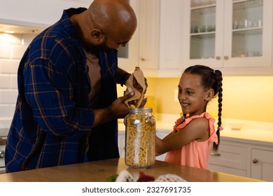 Happy biracial father and daughter pouring pasta from paper bag into storage jar in kitchen. Food, shopping, healthy lifestyle, ecology, family and domestic life, unaltered. - Powered by Shutterstock