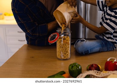 Happy biracial father and son pouring pasta from paper bag into storage jar in kitchen. Food, shopping, healthy lifestyle, ecology, family and domestic life, unaltered. - Powered by Shutterstock